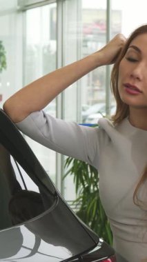 Akan kızıl saçlı ışıl ışıl genç bir kadın akıllı telefonuyla canlı bir sohbete dalarken çağdaş bir araba galerisinde zarif bir şekilde şık bir araca yaslanıyor. Aydınlık, sergi salonu, yemyeşil.