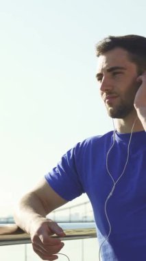 Düşük açılı bir görüntü, canlı mavi tişörtlü, yakışıklı, sakallı bir adamı enerji veren bir egzersizden sonra açık havada müzik dinlemekten zevk alırken mutlu bir şekilde gülümseyerek yakalar. Bu canlandırıcı sahne pozitiflik ve sükunet yayıyor, aktif bir ruhun somutlaştırılması.