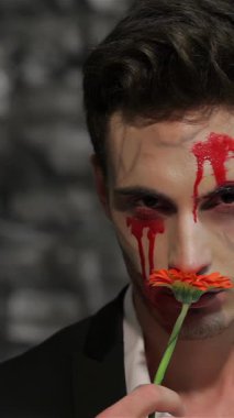 Gösterişli siyah takım elbiseli çarpıcı bir erkek vampir burnuna canlı bir turuncu çiçek getiriyor ve kanlı yüzüyle rahatsız edici bir kontrast oluşturuyor. Koyu, desenli tuğla bir duvara karşı dizilmiş bu büyüleyici görüntü ürkütücü cazibeyi harmanlıyor.