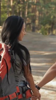 Neşeli bir çift yürüyüşçü, görkemli ağaçlar ve benekli güneş ışığıyla çevrili güneşli bir orman patikası boyunca el ele yürüyor. Çekici beyaz erkek ve kadın, açık hava maceralarını kucaklarken mutluluk saçıyor, aşkı somutlaştırıyor ve