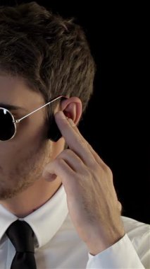 Keskin giyimli bir güvenlik görevlisi, spor gözlüklü, parlak bir kravat takmış, siyah bir arka plana karşı yoğun bir dikkatle etrafını tarıyor. Hareketleri ve ciddi davranışları otoriteyi ve hazırlığı yansıtıyor, özünü somutlaştırıyor.