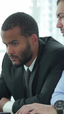 İki azimli iş adamı, biri şık takım elbiseli iyi bakımlı sakallı, diğeri de gevrek gömlekli, şık ve çağdaş bir ofis masasında yoğun bir şekilde iş birliği yapıyorlar. Ciddi ifadeleri ve ilgili vücut dilleri,