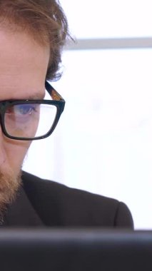 A sharply dressed Caucasian businessman with a well-groomed beard is intensely focused on his laptop, immersed in a sea of financial data. The soft backlight from the window casts a warm glow, accentuating his serious expression and highlighting his