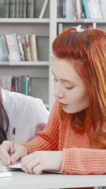 In a vibrant library brimming with colorful books, two female students are passionately collaborating on a project. One student, with striking red hair, diligently writes notes while her focused Asian friend reviews information on a sleek laptop