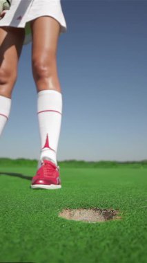 Kırmızı golf ayakkabılarına odaklanmış genç bir kadın topu fincana doğru atarken kararlılığını belli bir vuruşla ustalıkla ortaya koyuyor. Bereketli yeşil golf sahası, parlak güneş ışığı altında parıldıyor.