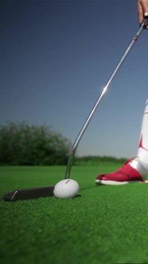 A focused young woman in striking red golf shoes expertly holes out with a precise putt, her determination shining through as she skillfully strikes the ball towards the cup. The lush green golf course sparkles under the radiant sunlight, creating an