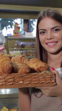 In a cozy bakery, a charming young woman in a stylish apron beams with delight as she holds a wicker basket filled with golden, flaky croissants. Her radiant smile and playful finger-kissing gesture capture the essence of culinary joy, inviting