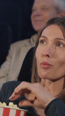 An attractive Caucasian woman, with long flowing hair, playfully pops popcorn into her mouth, her eyes sparkling with excitement as she immerses herself in the cinematic experience. Surrounded by a lively audience, her infectious laughter and radiant