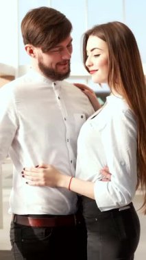 A stylish brunette man excitedly gestures towards a sleek car, while his elegant girlfriend leans in, sharing a moment of joy in the bright, modern dealership. The inviting atmosphere is filled with a variety of vehicles, making their car-buying