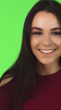 A close-up of a radiant young woman with long brunette hair, joyfully smiling at the camera while forming a heart shape with her hands against a vibrant green backdrop. This lively studio shot captures her playful spirit and the essence of love and