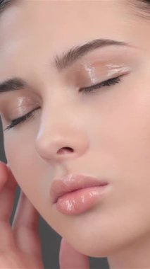 This exquisite close-up captures a young brunette woman gently tilting her head, eyes softly closed in a blissful moment of tranquility. As she tenderly massages her temple and cheek, her flawless skin glows against a soothing gray backdrop