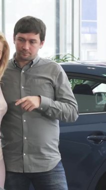 A mature couple stands together in a sleek, modern dealership, animatedly discussing their options for a new automobile. The man points enthusiastically at a stunning blue car, while his radiant wife beams with joy, embodying excitement as they