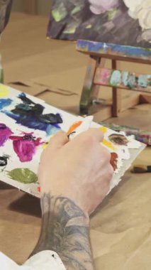 In this intimate close-up, a male painter passionately squeezes vibrant oil paint onto his palette, surrounded by an array of brushes and a blank canvas. The warm, inviting atmosphere of his cozy art studio radiates creativity and inspiration