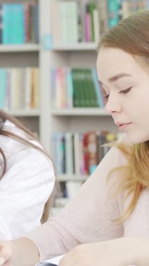Two dedicated teenage girls are deeply engaged in their studies at a lively library, collaborating on an assignment with a laptop and colorful notebooks. Their focused expressions radiate camaraderie and determination, capturing the essence of