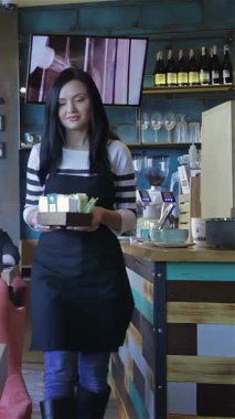 A vibrant young waitress with a beaming smile stands confidently at the counter of a cozy cafe, ready to take orders. Dressed in a stylish black apron and a striped shirt, she embodies warmth and hospitality, surrounded by the rich aroma of freshly