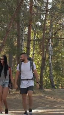 A delightful couple of hikers strolls hand in hand along a sun-drenched forest path, radiating happiness and connection. The brunette woman playfully adjusts her hair while the bearded man shares a warm smile, capturing the essence of adventure and