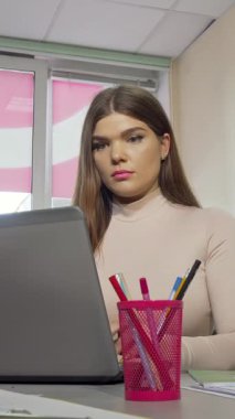 A weary female student with long brown hair and striking nails rubs her temples in frustration as she intensely types on her laptop, surrounded by vibrant stationery in a bright, modern workspace. This scene vividly illustrates the emotional strain