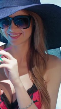 Canlı kırmızı mayo giymiş çarpıcı derecede zayıf bir kadın tropikal bir kokteyl yudumluyor. Koyu mavi şapkası parlak gülüşünü vurguluyor. Işıl ışıl havuz suyu güneşin altında parıldıyor, bu mutlu yaz kaçışı için huzurlu bir ortam yaratıyor.