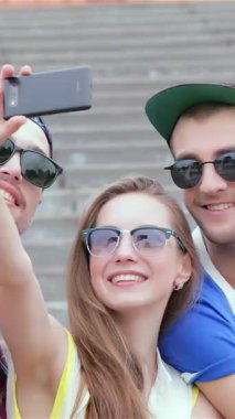 Canlı bir arkadaş üçlüsü, sıcak güneş ışığında güneşleniyor, merdivenlerde neşeyle selfie çekiyor, mutluluk ve dostluk saçıyor. Şık güneş gözlükleri ve renkli kıyafetleri bu anı gerçekten unutulmaz kılacak canlı bir yaz atmosferi yaratıyor.