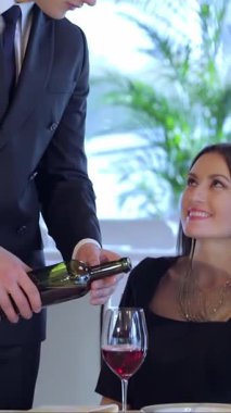 Şık bir genç çift zarif bir restoranda yıldönümü kutlamalarında eğlenirken, koca eşi için bir bardak kadife kırmızı şarap dolduruyor. Mum ışığının yumuşak parıltısı ve yemyeşil yeşillik onları sarıp sarmalayarak samimi bir atmosfer yaratıyor.