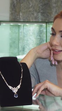 Pürüzsüz, siyah bir standın üzerinde zarif bir şekilde sergilenen nefes kesici elmas kolyeye hayranlık duyan, neşe saçlarıyla ışıldayan genç bir kadın. Pırıl pırıl mücevherler ve hafif ışıklandırma ile süslenmiş şık kuyumcunun lüks atmosferi.