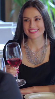 Zarif giysiler ve pırıl pırıl mücevherlerle süslenmiş göz kamaştırıcı genç bir kadın kadeh kaldırarak kadeh kaldırırken neşe saçıyor. Yumuşak mum ışığında yıkanan ve yemyeşil yemyeşil ile çevrili samimi restoran ortamı büyülü bir şey yaratır.