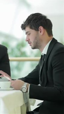 A sharp-dressed young man, with dark hair and a focused expression, intently reviews information on his tablet while seated at a sleek, modern table. The bright, airy office setting enhances the atmosphere of collaboration and innovation, as