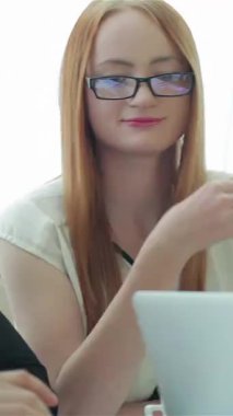 A focused woman with long red hair and glasses engages in a dynamic discussion with a businessman, surrounded by important documents in a modern office. Their interaction exudes confidence and collaboration, creating an inspiring atmosphere of