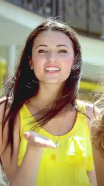 Three stylish young women, radiating joy, blow playful kisses amidst a bustling shopping district, their colorful bags swinging in the warm sunlight. Their laughter dances through the air, creating an infectious atmosphere of friendship and carefree