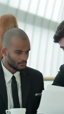 Two confident businessmen, one with a shaved head and the other with dark hair, are intensely reviewing documents at a modern office table, exuding an air of professionalism and collaboration. Their expressions reflect determination and success