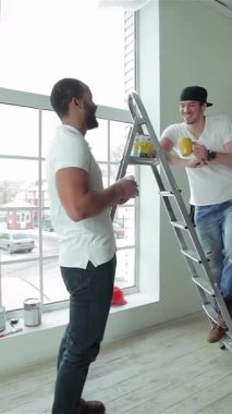 Two cheerful workers share a light-hearted snack break in a sunlit, freshly renovated apartment, radiating joy and camaraderie. One man, perched on a sturdy ladder, hands paint to his colleague, highlighting their teamwork in this modern, airy space