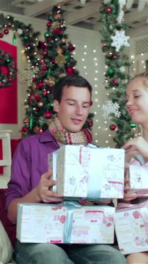 A cheerful couple, bundled in cozy sweaters and scarves, joyfully unwraps beautifully decorated gifts in a magical Christmas room filled with twinkling lights and a stunning tree. This heartwarming scene radiates love and holiday spirit, capturing