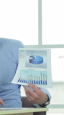In a bright, modern office, two confident businessmen engage in a dynamic discussion, passionately analyzing a detailed business plan. One man, in a crisp light blue shirt, holds up colorful charts, while the other, dressed in a sleek black suit