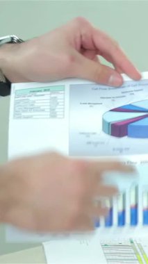 A close-up showcases two businessmen animatedly discussing colorful graphs on a business report, their hands gesturing with enthusiasm. Clad in sharp formal attire, they radiate confidence and professionalism in a sleek, modern office filled with