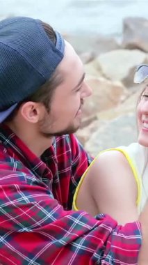 A radiant couple sits on a sun-kissed rocky shore, playfully touching foreheads and exchanging warm smiles. Their laughter dances in the air, embodying a blissful summer day filled with love, joy, and connection