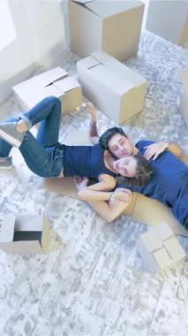 In a bright, sun-drenched room filled with unpacked cardboard boxes, a couple lies on the floor in a loving embrace, exuding happiness and excitement. Their laughter fills the air, marking the start of a beautiful journey together in their new home