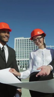 Three confident business architects, a man and two women in bright orange helmets, stand outdoors under a clear blue sky, eagerly examining blueprints. Their dynamic teamwork and shared vision exude excitement and innovation as they passionately