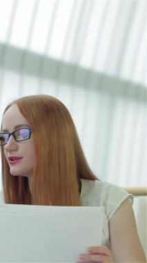 Kendine güvenen kızıl saçlı ve şık gözlükleri olan bir kadın, her ikisi de önemli belgeleri incelemeye dalmış iyi giyimli bir adamla tutkuyla fikir alışverişinde bulunuyor. Arka planda, diğer iki profesyonel, bir erkek ve bir kadın, dikkatle