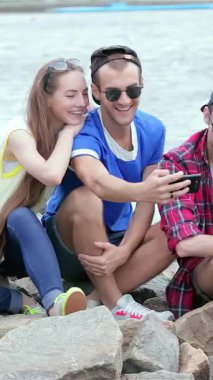Üç genç arkadaş, iki erkek ve bir kadın, sıcak güneşin altında neşe ve dostluk saçan kayalık bir nehir kenarında selfie çekiyorlar. Kahkahaları ve enerjik enerjileri bu anı mükemmel bir enstantane haline getirecek canlı bir atmosfer yaratıyor.