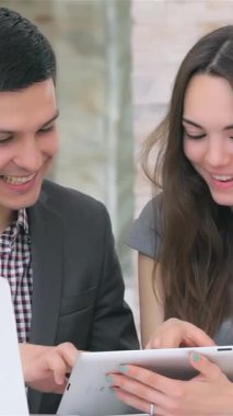 Genç, çekici bir iş kadını ve işadamı, modern bir tablet üzerindeki verileri hevesle tartışarak dinamik bir işbirliği anını paylaşıyorlar. Onların parlak gülümsemeleri ve odaklanmış ifadeleri çağdaş ofisi aydınlatıyor, doğal ışık ve