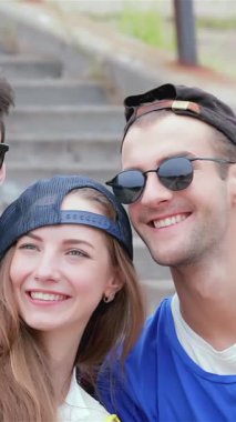 Neşeyle ışıldayan canlı bir grup genç arkadaş, açık hava basamaklarında neşeli bir poz vererek akıllı telefonlarıyla selfie çekiyorlar. Şık şapkaları ve güneş gözlükleri güneşli atmosferi tamamlar. Kaygısız yaz atmosferleri yayar.