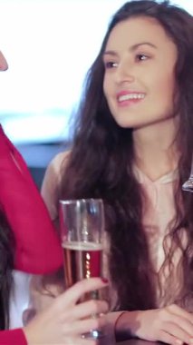 İki neşeli genç kadın, biri uzun siyah saçlı, diğeri dalgalı esmer saçlı, canlı bir barda arkadaşlıklarını kutluyor, kadehlerini neşeli bir şekilde kaldırıyorlar. Hayat dolu atmosfer kahkahalarla ve arkadaşlıkla dolu.