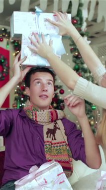 Peri ışıklarıyla süslenmiş sıcacık bir Noel odasında ve büyüleyici dekore edilmiş bir ağaçla, neşeli bir çift birbirlerine güzel paketlenmiş hediyeler veriyor, yüzleri neşe ve şaşkınlık saçıyor. Tatil neşesiyle dolu sıcak atmosfer,