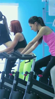 Canlı bir spor salonunda parlak mavi duvarlarla süslenmiş genç bir kız, kendini adamış arkadaşları sabit bisikletler üzerinde yoğun bir bisiklet egzersizi yaparken ferahlatıcı bir yudum su alır. Bu dinamik sahne enerji ve yoldaşlık yayar.