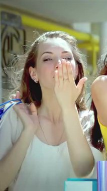 Üç şık genç kadın, her biri canlı alışveriş torbaları tutuyor, kameraya şakacı öpücükler saçıyor, neşe ve dostluk saçıyor. Işıl ışıl gülümsemeleri ve renkli kıyafetleri canlı, güneşli bir atmosfer yaratıyor.