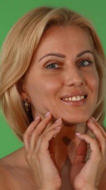 Işıl ışıl sarı saçları ve çarpıcı mavi gözleri olan olgun bir kadın kameraya sıcak bir şekilde gülümsüyor, yanağını canlı yeşil bir kromakey arka planına hafifçe okşuyor. Bu büyüleyici görüntü güzelliği, özgüveni ve sağlıklı bir güzelliği kutluyor.