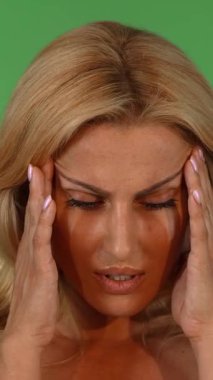 Canlı, yeşil bir krom anahtar stüdyosunda, dalgalı sarı saçlı, çarpıcı olgun bir kadın şakaklarını nazikçe ovuyor, ifadesi baş ağrısının ağırlığını yansıtıyor. Zarif özellikleri ve katlandığı rahatsızlıklar arasındaki zıtlık...