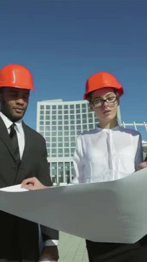 Kendine güvenen iş mimarlarından oluşan dinamik bir ikili, şık takım elbiseler ve canlı turuncu miğferler giymiş bir erkek ve bir kadın, parlak mavi gökyüzünün altındaki planları titizlikle inceliyor. Odaklanmış ifadeleri ve işbirlikçi enerjileri,