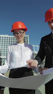 Üç kendine güvenen mimar, şık takımlar ve parlak turuncu kasklar giyip, açık havada detaylı bir iş planını tutkuyla gözden geçiriyorlar. Odaklanmış ifadeleri ve işbirlikçi enerjileri, yenilikçi fikirleri dönüştürmeyi tasarladıkları için heyecan yayıyor.