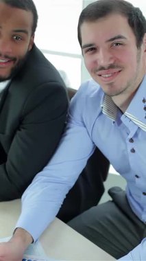 Dinamik bir iş ikilisi, parlak siyah takım elbiseli bir adam ve açık mavi gömlekli bir adam, neşeli bir şekilde parmaklarını şık çizelgeler ve bir dizüstü bilgisayarla dolu şık bir masada sergiliyorlar. Işıl ışıl gülüşleri ve kendinden emin tavırları,